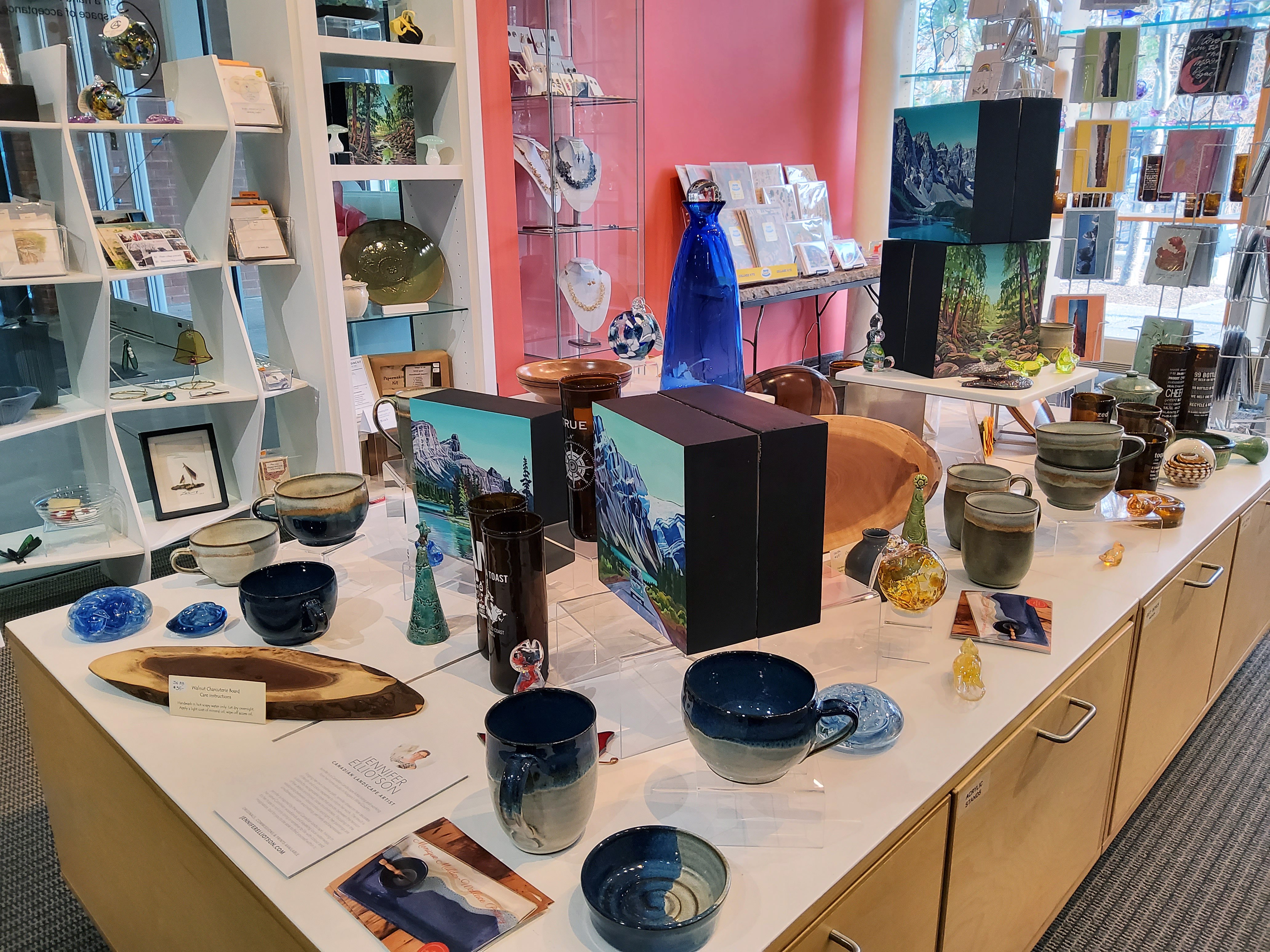 white shelving and display tables displaying various handmade items including pottery, handblown glass, greeting cards, jewellery, handcarved wood bowls and more