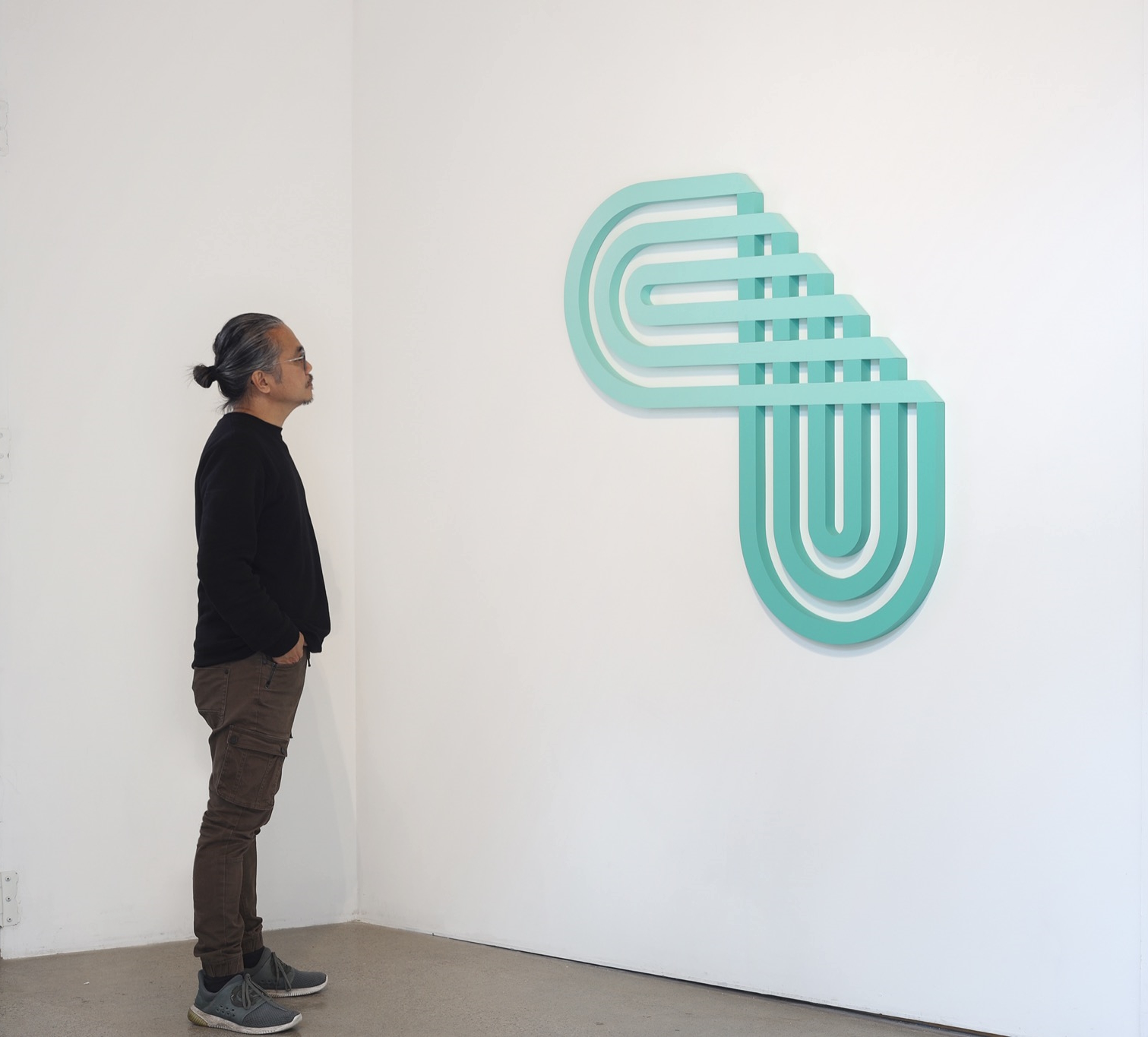 a man looking at a turquoise abstract 2D sculpture hanging on a white wall